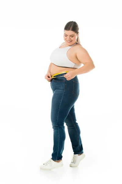 Улыбающаяся женщина с избыточным весом в джинсах, измеряющая талию с измерительной лентой, изолированной на белом, концепция позитивности тела — стоковое фото