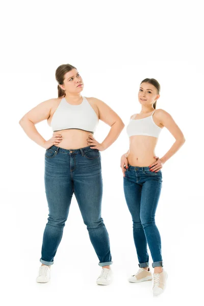 Alegres mujeres delgadas y tristes con sobrepeso en denim posando con las manos en las caderas juntas aisladas en blanco, concepto de positividad corporal - foto de stock