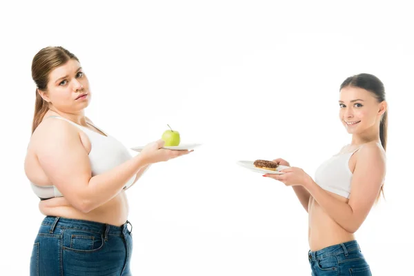 Felice donna magra e donna in sovrappeso in posa insieme con ciambella e mela verde su piatti isolati su bianco — Foto stock