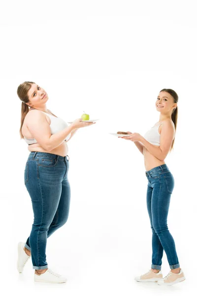 Donna magra e donna sovrappeso in posa insieme a ciambella e mela verde su piatti isolati su bianco — Foto stock