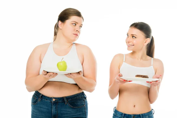 Felice donna magra che tiene piatto con ciambella e guardando triste donna in sovrappeso con mela verde sul piatto isolato su bianco — Foto stock