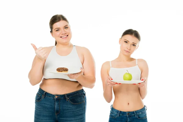 Sad slim woman holding plate with green apple and happy overweight woman holding plate with doughnut and showing thumb up isolated on white — Stock Photo