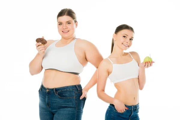 Счастливые стройные и толстые девушки позируют вместе с пончиком и зеленым яблоком изолированы на белом — стоковое фото