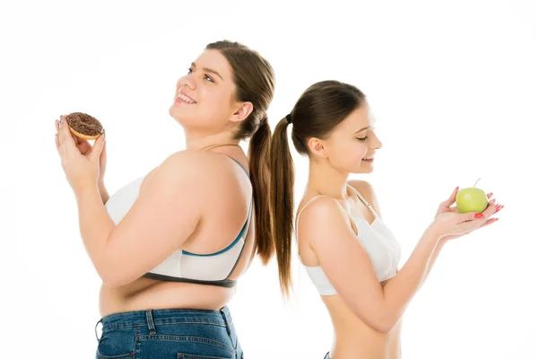 Felici ragazze magre e sovrappeso in piedi schiena contro schiena insieme con ciambella e mela verde isolato su bianco — Foto stock