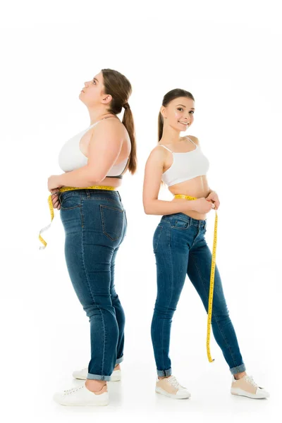 Triste mujer con sobrepeso con cinta métrica mirando hacia arriba mientras atractiva mujer delgada mirando a la cámara aislada en blanco - foto de stock