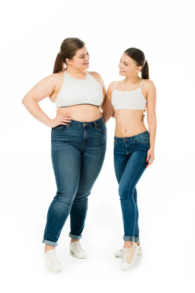 Mujeres delgadas y con sobrepeso felices abrazándose y mirándose aislados en blanco, concepto de positividad corporal - foto de stock