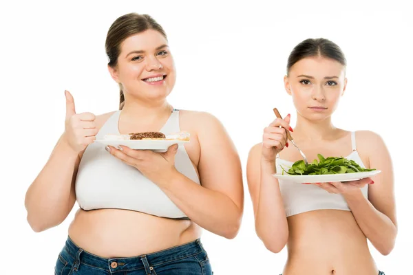 Happy overweight woman with doughnuts showing thumb up while sad slim woman holding plate with green spinach leaves isolated on white — Stock Photo