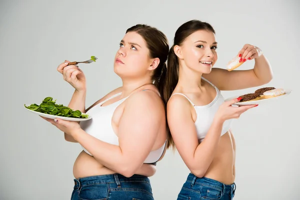 Glückliche schlanke Frau isst Donuts, während traurige übergewichtige Frau grüne Spinatblätter isst — Stockfoto
