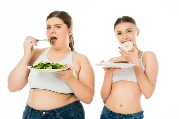 Donna magra mangiare ciambelle mentre donna in sovrappeso mangiare foglie di spinaci verdi isolate su bianco — Foto stock