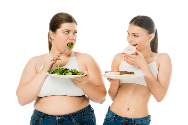 Schlanke Frau isst Krapfen und übergewichtige Frau isst grüne Spinatblätter, während sie sich isoliert auf weißem Grund ansieht — Stockfoto
