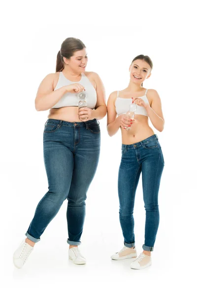 Mulheres felizes magras e com excesso de peso em jeans segurando garrafas com água isolada em branco — Fotografia de Stock