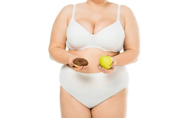 Обрезанный вид женщины с избыточным весом в нижнем белье с пончиком и зеленым яблоком, изолированным на белом — стоковое фото
