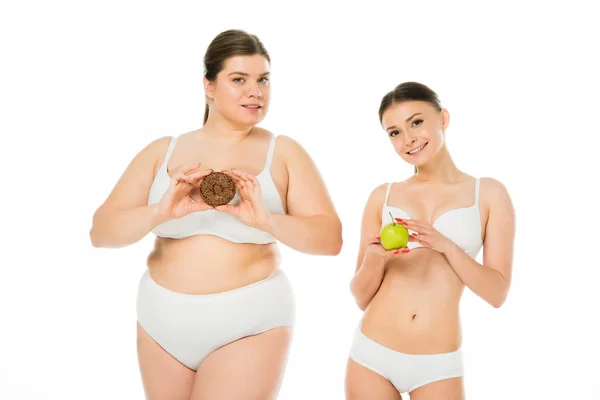 Joven mujer delgada sosteniendo manzana verde mientras mujer con sobrepeso sosteniendo donut dulce aislado en blanco - foto de stock