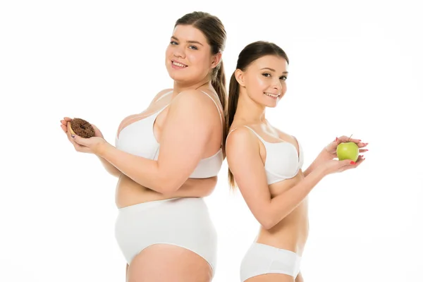 Junge schlanke Frau mit grünem Apfel, Rücken an Rücken stehend mit übergewichtiger Frau mit süßem Donut, isoliert auf weiß — Stockfoto