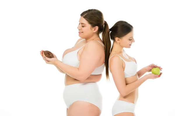 Happy slim woman with green apple standing back to back with smiling overweight woman with sweet doughnut isolated on white — Stock Photo