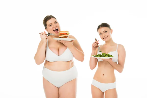 Feliz mujer delgada de pie con plato de hojas de espinacas verdes mientras sonríe mujer con sobrepeso comiendo hamburguesa aislada en blanco - foto de stock