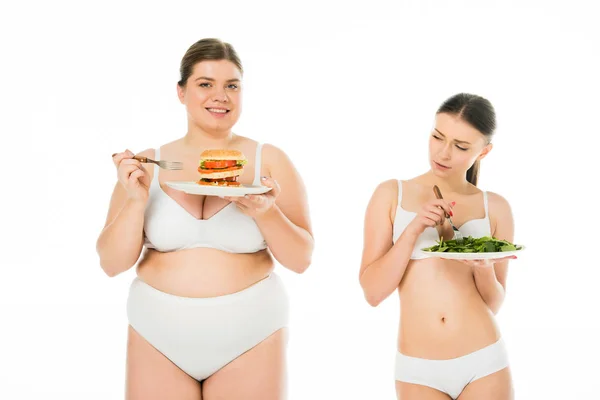 Disgustada mujer delgada de pie con plato de hojas de espinacas verdes mientras sonríe mujer con sobrepeso sosteniendo plato con hamburguesa aislada en blanco - foto de stock