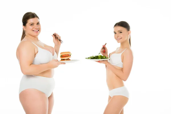 Mujer delgada en ropa interior de pie con placa de hojas de espinaca verde mientras que la mujer con sobrepeso sostiene la placa con hamburguesa aislada en blanco - foto de stock