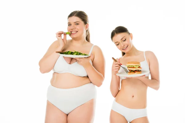 Mujer delgada en ropa interior mirando hamburguesa mientras que la mujer con sobrepeso comiendo hojas de espinacas verdes aisladas en blanco - foto de stock