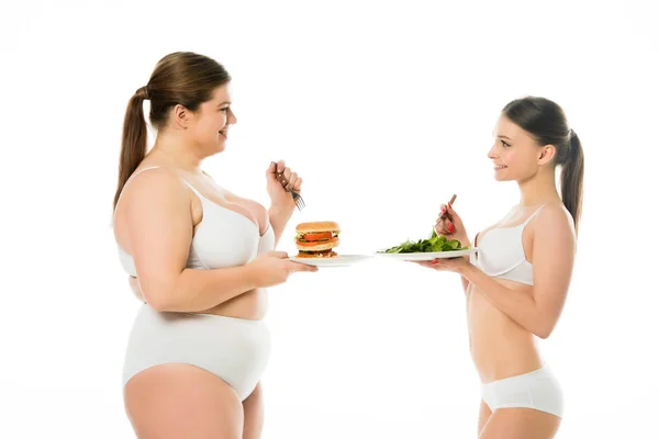 Schlanke Frau in Unterwäsche mit Teller mit grünen Spinatblättern und Blick auf übergewichtige Frau mit Teller mit Burger isoliert auf weiß — Stockfoto