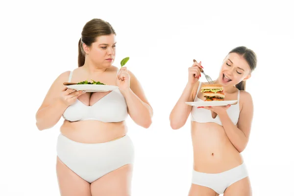 Donna magra in biancheria intima mangiare hamburger mentre donna sconvolta in sovrappeso tenendo foglia di spinaci verde isolato su bianco — Foto stock