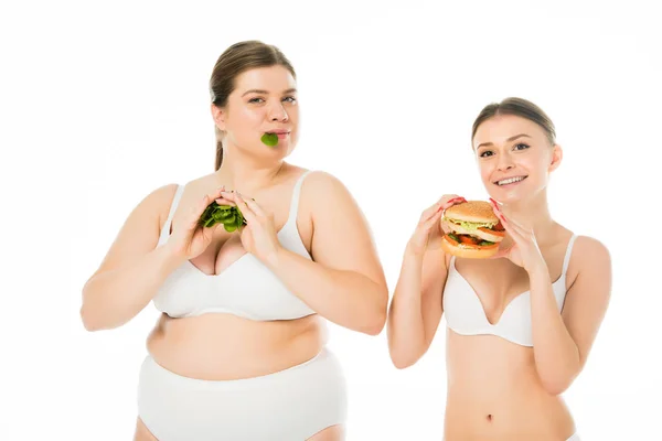 Delgada mujer sonriente en ropa interior sosteniendo hamburguesa mientras que la mujer feliz con sobrepeso comiendo hojas de espinacas verdes aisladas en blanco - foto de stock