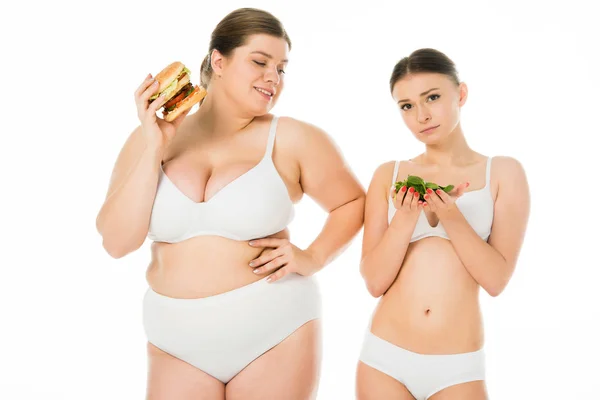 Sottile donna sconvolta in biancheria intima con foglie di spinaci verdi mentre in sovrappeso donna felice mangiare hamburger isolato su bianco — Foto stock