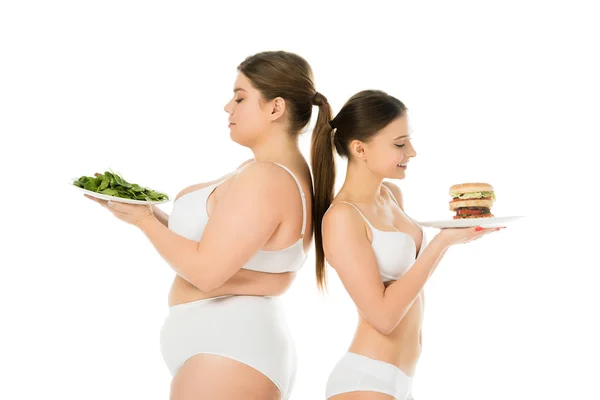 Felice donna magra con hamburger in piedi schiena a schiena con donna sovrappeso sconvolto con foglie di spinaci verdi isolate su bianco — Foto stock