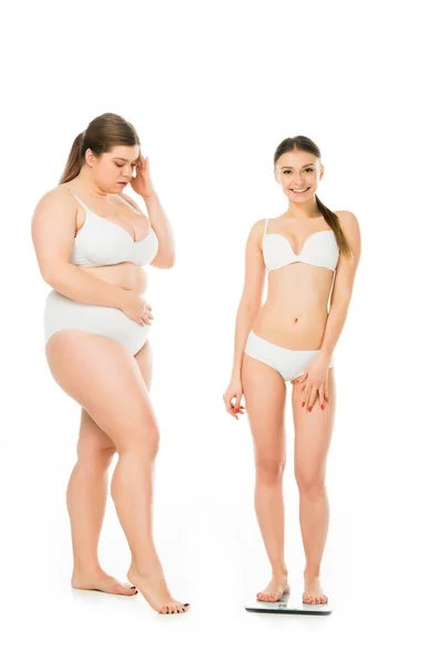 Impactado mujer con sobrepeso en ropa interior mirando feliz mujer delgada en escamas aisladas en blanco, concepto de positividad corporal - foto de stock