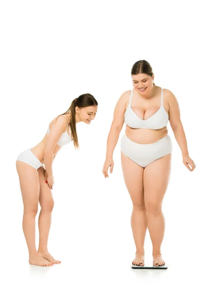 Lächelnde schlanke Frau in Unterwäsche, die eine glückliche übergewichtige Frau auf einer Waage sieht, die auf einer weißen Waage isoliert ist, Konzept der Körperpositivität — Stockfoto