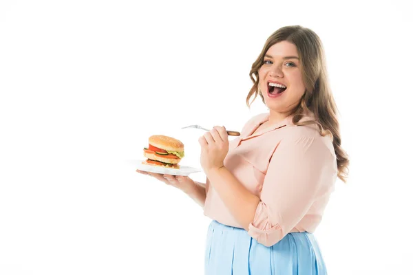 Щаслива усміхнена жінка з відкритим ротом тримає смачний бургер на тарілці і виделці ізольовані на білому — стокове фото