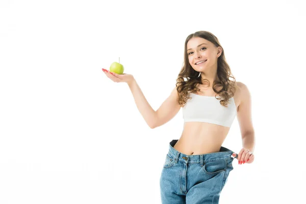 Smiling slim young woman in big jeans holding green apple isolated on white — Stock Photo