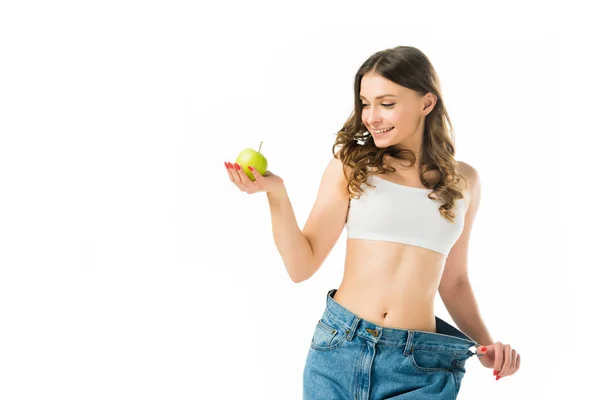 Happy slim young woman in big jeans holding green apple isolated on white — Stock Photo