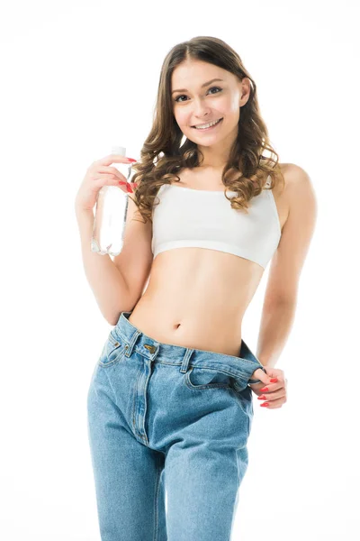 Усміхнена струнка дівчина тримає у великих джинсах пляшку з водою ізольовано на білому, схуднути концепція — стокове фото
