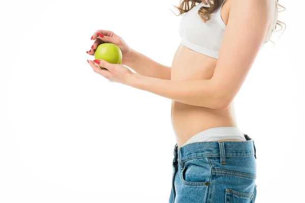 Обрізаний вид тонкої молодої жінки у великих джинсах, що тримає зелене яблуко, ізольоване на білому — стокове фото