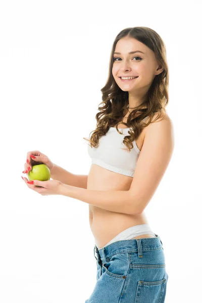 Вид збоку усміхненої стрункої молодої жінки у великих джинсах, що тримає зелене яблуко, ізольоване на білому — стокове фото