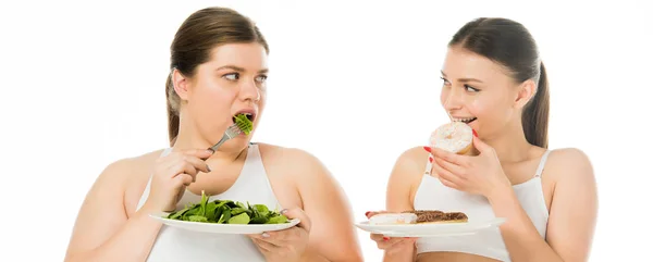 Mujer delgada comiendo donas y mirando a la mujer con sobrepeso comiendo hojas de espinacas verdes aisladas en blanco - foto de stock