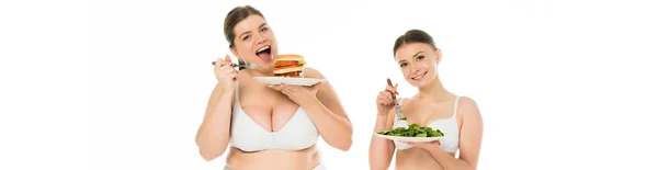 Sovrappeso donna sorridente in biancheria intima mangiare hamburger dal piatto mentre sottile donna felice mangiare foglie di spinaci verdi isolato su bianco — Foto stock
