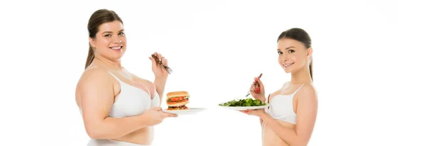 Femme souriante en surpoids en sous-vêtements tenant un hamburger sur une assiette tandis que la femme heureuse mince manger des feuilles d'épinards verts isolés sur blanc — Photo de stock