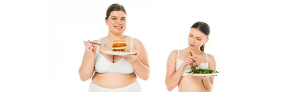 Femme souriante en surpoids en sous-vêtements tenant un hamburger sur une assiette tandis que la femme heureuse mince manger des feuilles d'épinards verts isolés sur blanc — Photo de stock