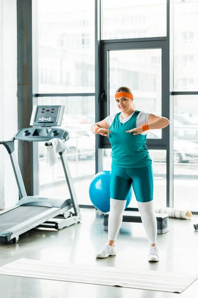 Übergewichtige Frau dehnt sich im Stehen in Sportbekleidung in der Nähe von Fitnessmatten — Stockfoto