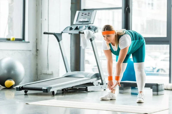 Übergewichtige Frau dehnt sich in der Nähe von Fitnessmatte und Laufband im Fitnessstudio — Stockfoto
