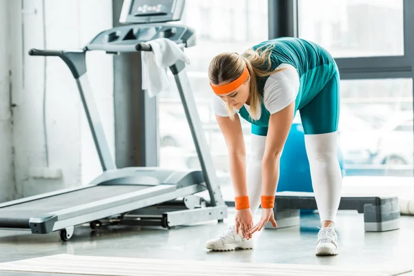 Concentrated overweight woman stretching near fitness mat and treadmill in gym — Stock Photo