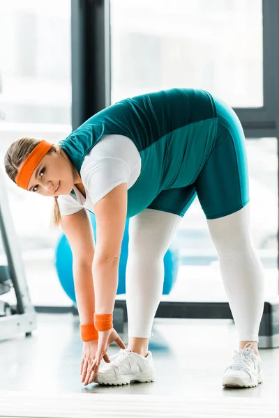 Overweight woman stretching near fitness mat and looking at camera — Stock Photo