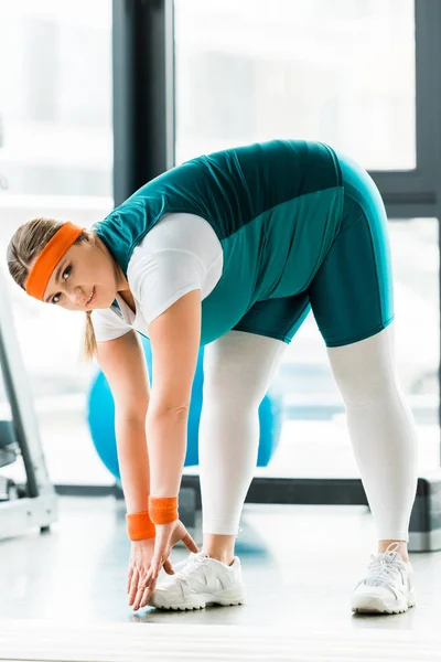 Mujer con sobrepeso estiramiento cerca de la estera de fitness y mirando a la cámara - foto de stock