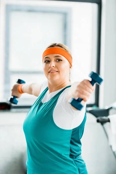 Selective focus of cheerful overweight woman workouting in sportswear with dumbbells — Stock Photo
