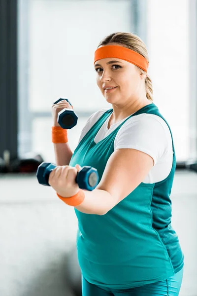 Избирательный фокус привлекательной толстой женщины, тренирующейся в спортивной одежде с гантелями — стоковое фото