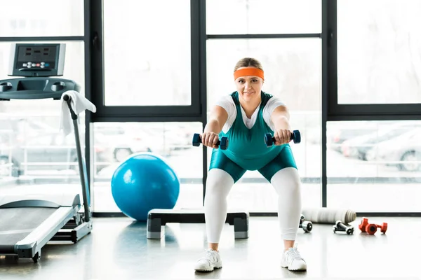 Femme en surpoids gaie séance d'entraînement avec haltères près du tapis roulant et balle de remise en forme dans la salle de gym — Photo de stock