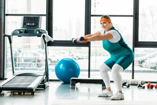Cheerful plus size woman exercising with dumbbells near treadmill — Stock Photo