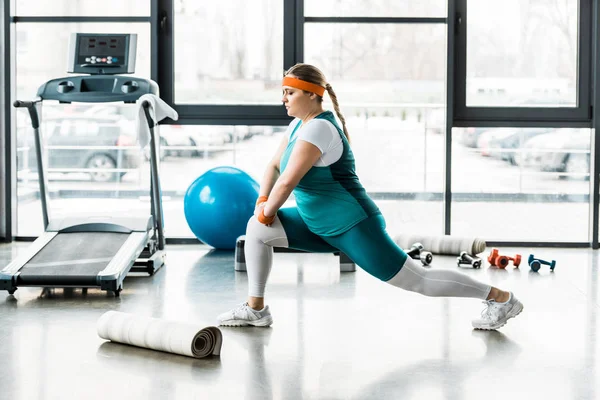 Confident plus size woman in sportswear stretching near fitness mat and treadmill — Stock Photo